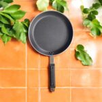 Non-stick 25 cm black tapered fry pan with a long handle, placed on a warm terracotta tile surface surrounded by lush green indoor plants.