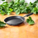 Non-stick 25 cm black tapered fry pan with a long handle, placed on a warm terracotta tile surface surrounded by lush green indoor plants.