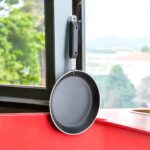 Non-stick 25 cm black tapered fry pan with a long handle, placed on a warm terracotta tile surface surrounded by lush green indoor plants.