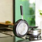 Non-stick 25 cm black tapered fry pan with a long handle, placed on a warm terracotta tile surface surrounded by lush green indoor plants.