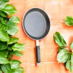 Non-stick 25 cm black tapered fry pan with a long handle, placed on a warm terracotta tile surface surrounded by lush green indoor plants.