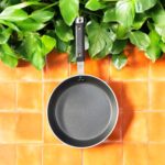 Non-stick 25 cm black tapered fry pan with a long handle, placed on a warm terracotta tile surface surrounded by lush green indoor plants.