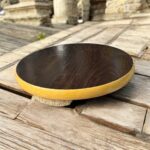 A round hardboard chakla with a dark brown top surface and yellow edges, featuring three anti-slip base supports, placed on a rustic wooden surface.