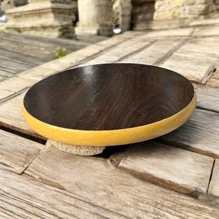 A round hardboard chakla with a dark brown top surface and yellow edges, featuring three anti-slip base supports, placed on a rustic wooden surface.