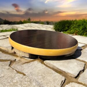 A round hardboard chakla with a dark brown top surface and yellow edges, featuring three anti-slip base supports, placed on a rustic wooden surface.