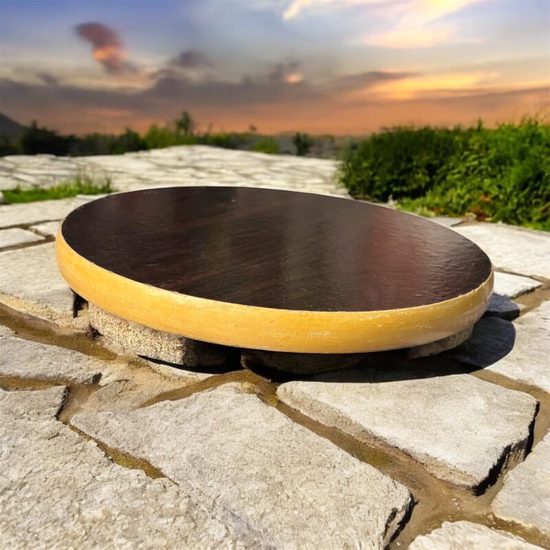 A round hardboard chakla with a dark brown top surface and yellow edges, featuring three anti-slip base supports, placed on a rustic wooden surface.