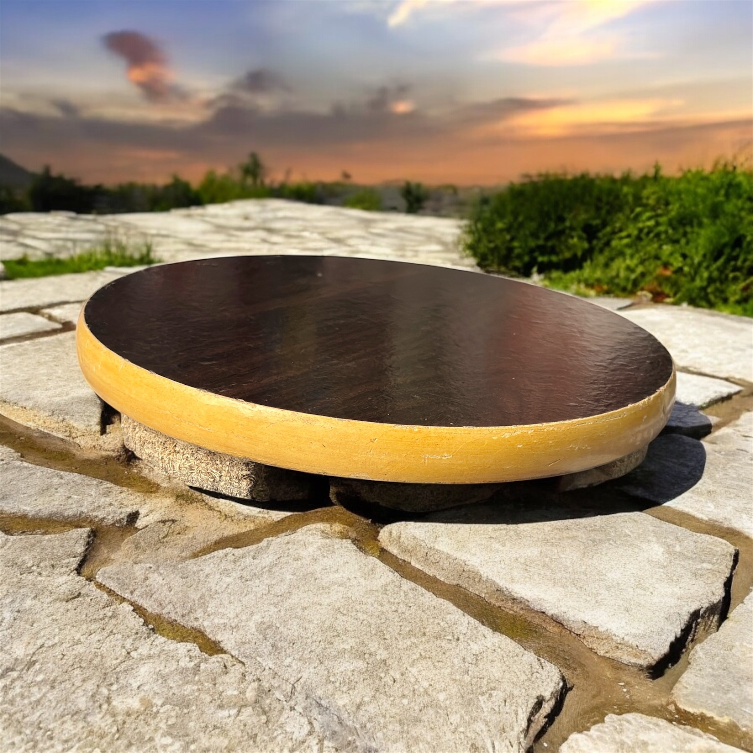 A round hardboard chakla with a dark brown top surface and yellow edges, featuring three anti-slip base supports, placed on a rustic wooden surface.