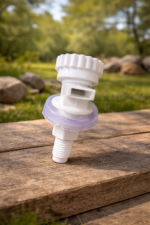 A white plastic water dispenser tap with a translucent purple seal, resting on a weathered wooden surface. The background is a soft-focus view of green grass, trees, and stones in natural daylight.