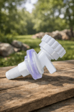 A white plastic water dispenser tap with a translucent purple seal, resting on a weathered wooden surface. The background is a soft-focus view of green grass, trees, and stones in natural daylight.