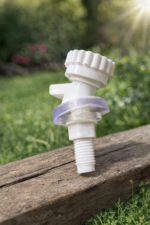 A white plastic water dispenser tap with a translucent purple seal, resting on a weathered wooden surface. The background is a soft-focus view of green grass, trees, and stones in natural daylight.