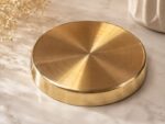 A highly polished, 7-inch Ruliarams pure Kansa (bronze) thali with a rich golden finish resting on a white marble surface, illuminated by natural sunlight with delicate dried flowers in the background.