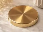 A highly polished, 7-inch Ruliarams pure Kansa (bronze) thali with a rich golden finish resting on a white marble surface, illuminated by natural sunlight with delicate dried flowers in the background.