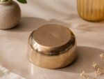 A highly polished, 5.5-inch Ruliarams pure Kansa (bronze) bowl featuring a gleaming metallic gold finish and hammered texture, resting on a warm, textured beige surface with soft natural shadows and delicate dried flowers in the background.
