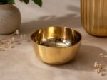 A highly polished, 5.5-inch Ruliarams pure Kansa (bronze) bowl featuring a gleaming metallic gold finish and hammered texture, resting on a warm, textured beige surface with soft natural shadows and delicate dried flowers in the background.