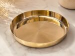 A highly polished, 7-inch Ruliarams pure Kansa (bronze) thali with a rich golden finish resting on a white marble surface, illuminated by natural sunlight with delicate dried flowers in the background.