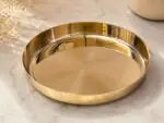 A highly polished, 7-inch Ruliarams pure Kansa (bronze) thali with a rich golden finish resting on a white marble surface, illuminated by natural sunlight with delicate dried flowers in the background.