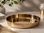 A highly polished, 7-inch Ruliarams pure Kansa (bronze) thali with a rich golden finish resting on a white marble surface, illuminated by natural sunlight with delicate dried flowers in the background.