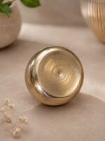 A highly polished, 5.5-inch Ruliarams pure Kansa (bronze) bowl featuring a gleaming metallic gold finish and hammered texture, resting on a warm, textured beige surface with soft natural shadows and delicate dried flowers in the background.